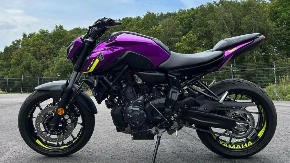 Lila Yamaha MT-07 motorcykel fotograferad i sidoprofil utomhus på asfalterad yta. Motorcykeln har svart ram och detaljer i neon-gult på fälgar och dekaler, med Yamaha-logotypen synlig på tanken och bakhjulet. I bakgrunden syns grön skog och stängsel, vilket ger en tydlig och sportig presentation av en custom- eller helfolierad motorcykel.